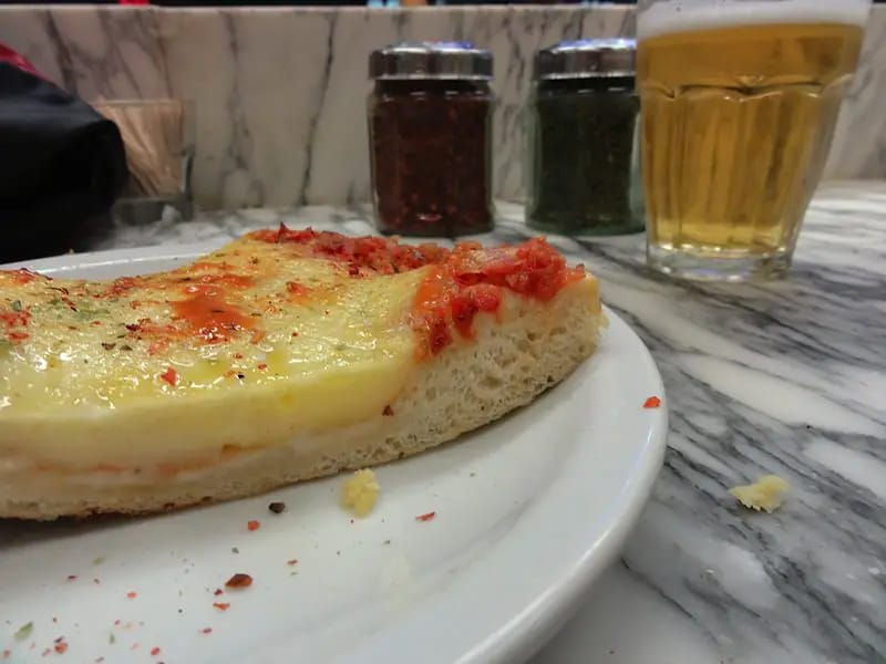 Thick Argentinian pizza with pepper, oregano and a beer behind.